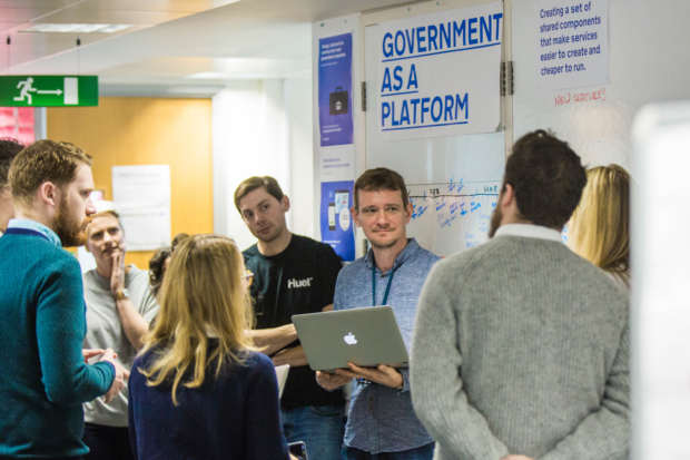 The government as a platform team stand up and discuss team matters in front of a sign that reads 'government as a platform'