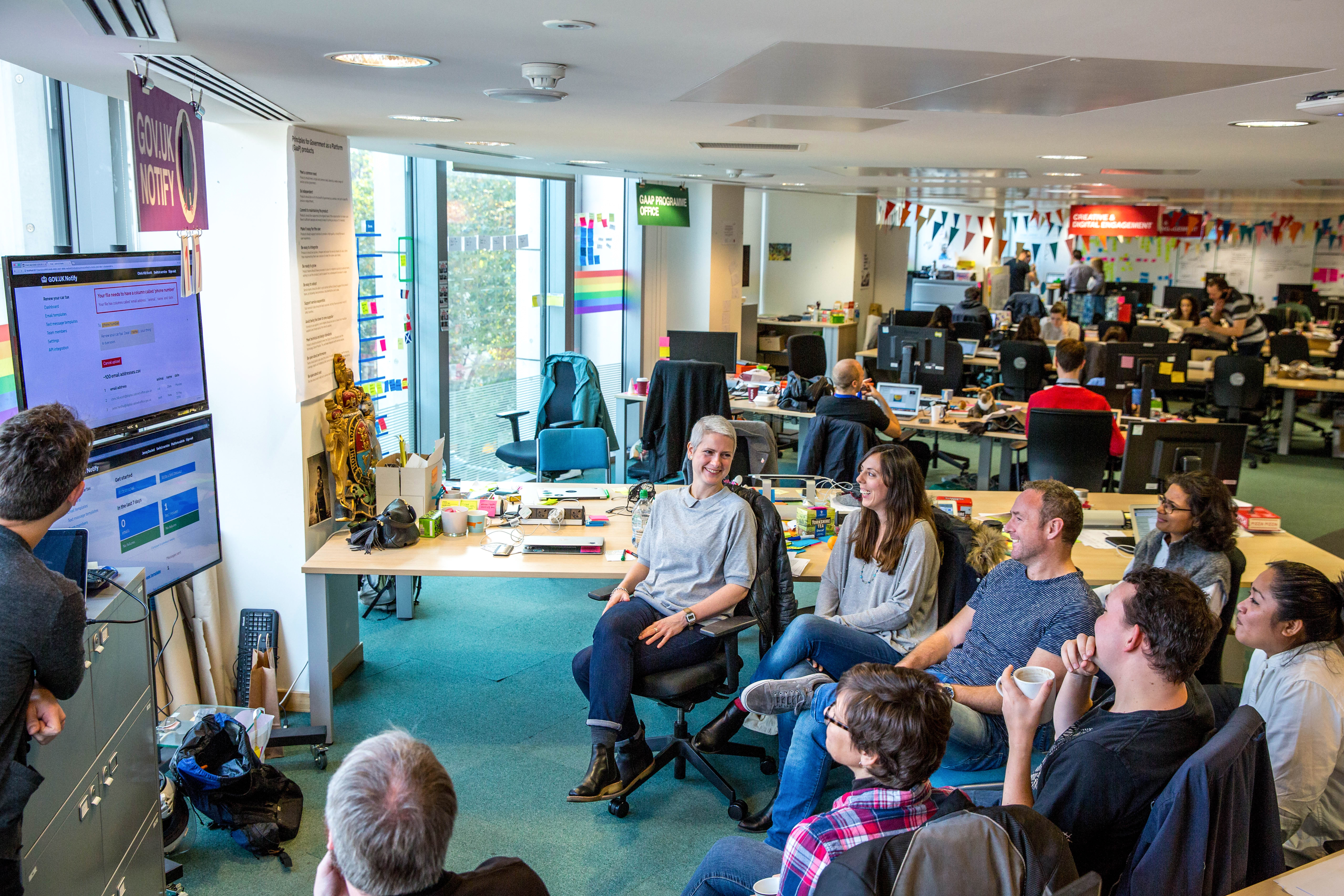 Photo of 9 members of the Notify team sitting in a semi circle in front of a screen showing the Notify dashboard.