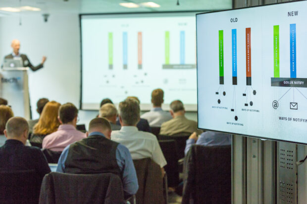 Photo of an event at which Government as a Platform components are showcased. The audience are facing screens showing a diagram of how GOV.UK Pay works